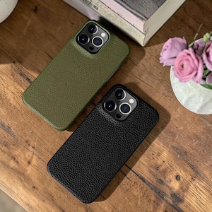 May include: Two phone cases, one olive green and one black, with a textured leather-like appearance. The cases are on a wooden surface, with a book and flowers in the background. The cases have multiple camera cutouts.