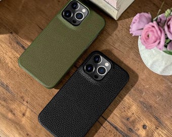 Puede incluir: Dos fundas para teléfono, una verde oliva y otra negra, con un aspecto texturizado similar al cuero. Las fundas están sobre una superficie de madera, con un libro y flores en el fondo. Las fundas tienen múltiples recortes para la cámara.