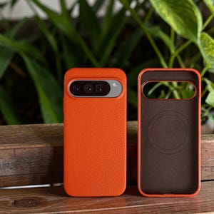 Puede incluir: Se muestran dos fundas de teléfono naranjas, una con un teléfono dentro y la otra vacía. Las fundas tienen una superficie texturizada y un interior marrón oscuro. La funda del teléfono de la izquierda tiene una barra de cámara plateada.