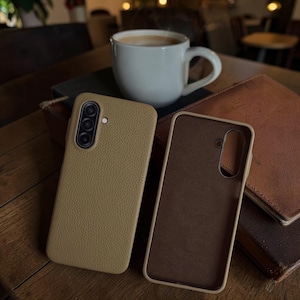 Op de afbeelding: Twee bruinleren telefoonhoesjes, &eacute;&eacute;n open, naast elkaar op een houten oppervlak. Een witte koffiemok staat op de achtergrond, samen met een stapel bruine leren notitieboekjes. De telefoonhoesjes hebben een textuur.