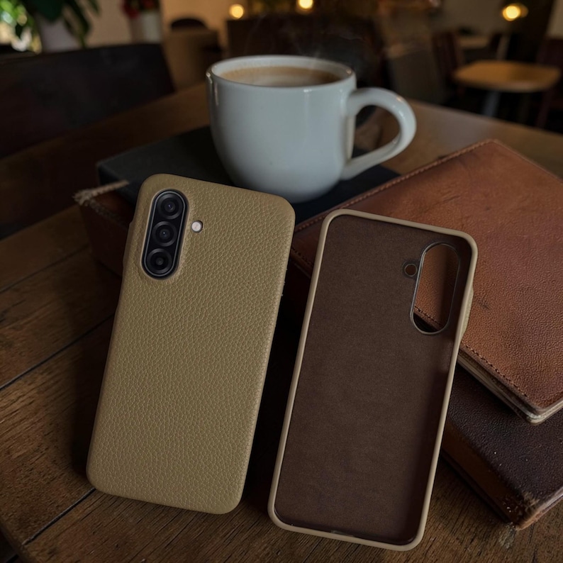 Op de afbeelding: Twee bruinleren telefoonhoesjes, &eacute;&eacute;n open, naast elkaar op een houten oppervlak. Een witte koffiemok staat op de achtergrond, samen met een stapel bruine leren notitieboekjes. De telefoonhoesjes hebben een textuur.