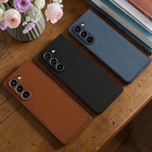 Puede incluir: Tres fundas texturizadas para tel&eacute;fono en marr&oacute;n, negro y azul se muestran sobre una superficie de madera. Cada funda tiene un m&oacute;dulo de c&aacute;mara con tres lentes circulares. Las fundas son rectangulares y parecen ser de cuero o material similar.