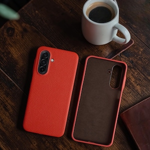 Op de afbeelding: Twee rode leren telefoonhoesjes, waarvan &eacute;&eacute;n open, liggen op een donker houten oppervlak. De hoesjes hebben een textuurafwerking en een camera-uitsparing. Een witte koffiemok met koffie en een klein leren notitieboekje liggen ook op de tafel.