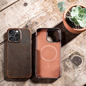 Pode incluir: Duas capas de telefone de couro marrom. Uma capa está montada, a outra aberta, revelando um interior marrom-rosado com um design circular. Elas estão em uma superfície de madeira, com uma pequena planta suculenta em vaso ao fundo.