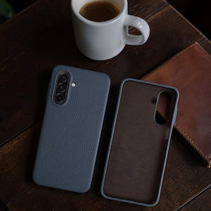 Op de afbeelding: Twee blauwe leren telefoonhoesjes, &eacute;&eacute;n open en &eacute;&eacute;n gesloten, liggen op een donker houten oppervlak. Een witte koffiemok en een bruine leren portemonnee staan ook op de tafel. De telefoonhoesjes hebben een getextureerde afwerking.