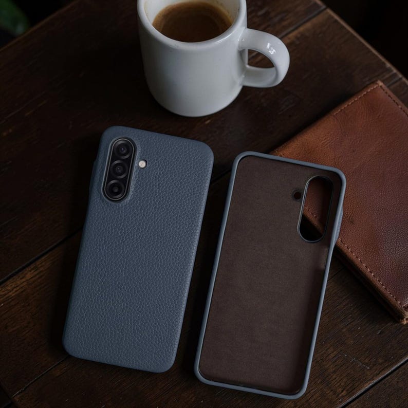 Op de afbeelding: Twee blauwe leren telefoonhoesjes, &eacute;&eacute;n open en &eacute;&eacute;n gesloten, liggen op een donker houten oppervlak. Een witte koffiemok en een bruine leren portemonnee staan ook op de tafel. De telefoonhoesjes hebben een getextureerde afwerking.