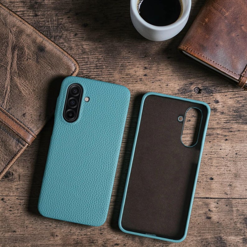 Op de afbeelding: Twee teal-gekleurde telefoonhoesjes, &eacute;&eacute;n open en &eacute;&eacute;n gesloten, met een textuur die op leer lijkt. De hoesjes liggen op een houten oppervlak, met een kopje koffie en een leren portemonnee op de achtergrond. De gesloten hoes heeft een cameramodule.