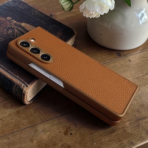 Puede incluir: Una funda de tel&eacute;fono de cuero marr&oacute;n con superficie texturizada. De forma rectangular, presenta un m&oacute;dulo de c&aacute;mara con tres lentes. Descansa sobre una superficie de madera junto a un libro antiguo.