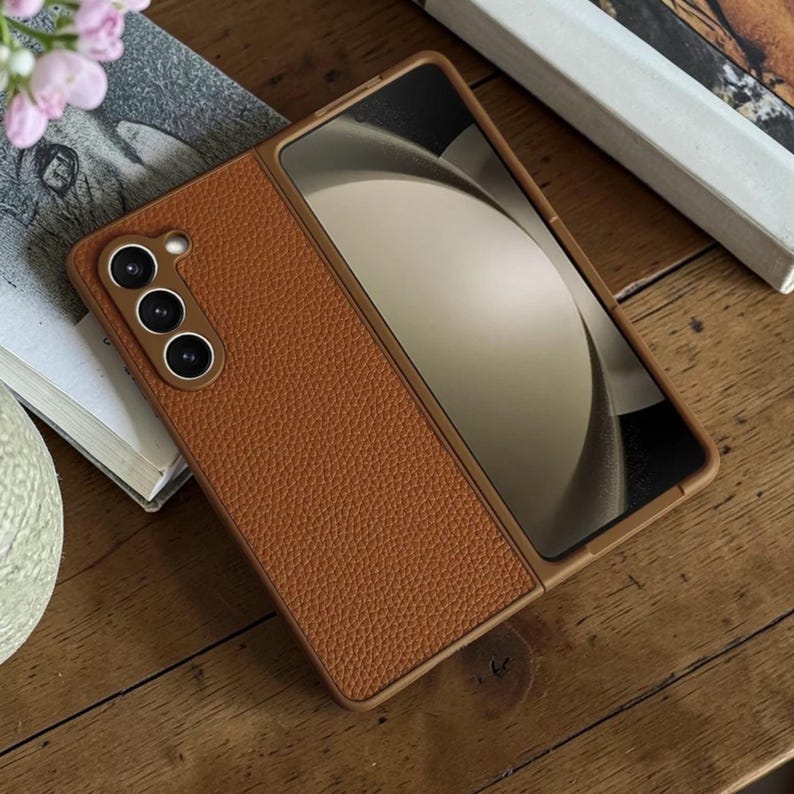 Puede incluir: Una funda de tel&eacute;fono de cuero marr&oacute;n con superficie texturizada. La funda est&aacute; abierta, mostrando la pantalla del tel&eacute;fono. Tiene un m&oacute;dulo de c&aacute;mara con tres lentes.