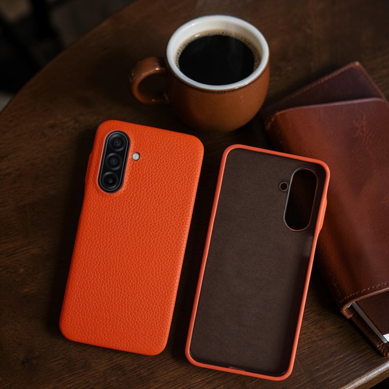 Op de afbeelding: Twee oranje leren telefoonhoesjes met een getextureerde afwerking worden gepresenteerd op een houten oppervlak. Een bruine koffiemok met zwarte koffie en een bruine leren portemonnee zijn ook aanwezig. De telefoonhoesjes hebben camera-uitsparingen.