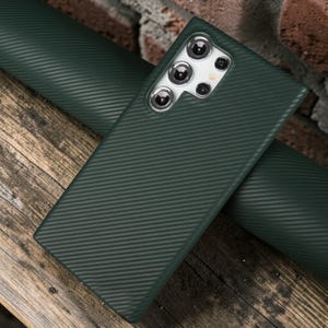 Könnte beinhalten: Dunkelgrüne Handyhülle mit Carbonfaserstruktur und mehreren Kameralinsen. Die Hülle liegt auf einer passenden grünen Materialrolle, mit einer Holzoberfläche im Hintergrund. Die Handyhülle dient zum Schutz des Telefons.