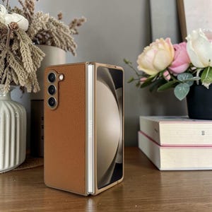 Puede incluir: Una funda de tel&eacute;fono de cuero marr&oacute;n para un tel&eacute;fono Samsung Galaxy Z Fold. La funda est&aacute; abierta, revelando la pantalla del tel&eacute;fono. La funda tiene una superficie texturizada y un marco dorado. El tel&eacute;fono tiene tres lentes.