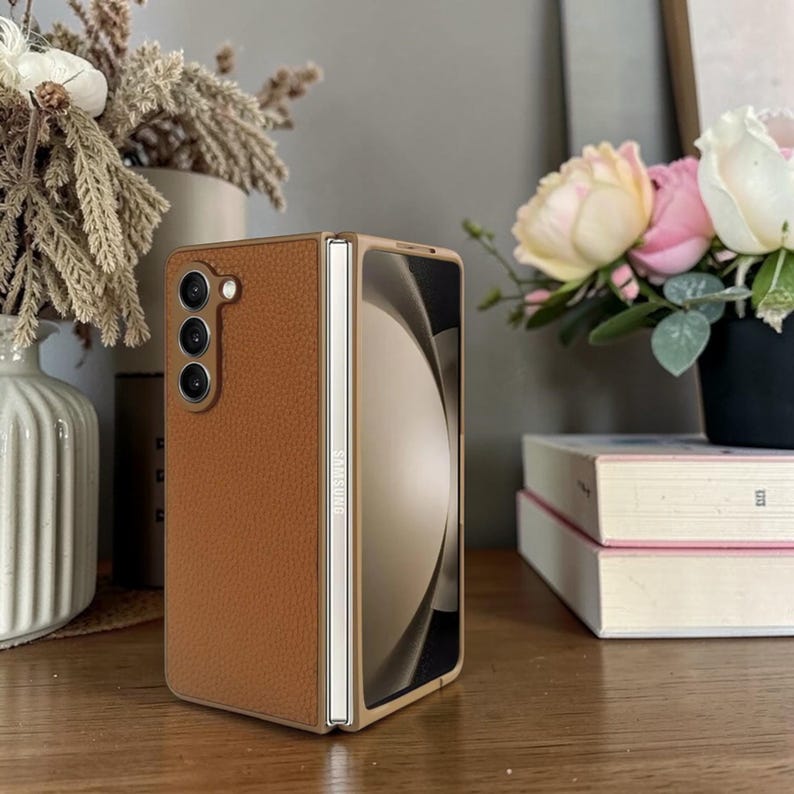 Puede incluir: Una funda de tel&eacute;fono de cuero marr&oacute;n para un tel&eacute;fono Samsung Galaxy Z Fold. La funda est&aacute; abierta, revelando la pantalla del tel&eacute;fono. La funda tiene una superficie texturizada y un marco dorado. El tel&eacute;fono tiene tres lentes.