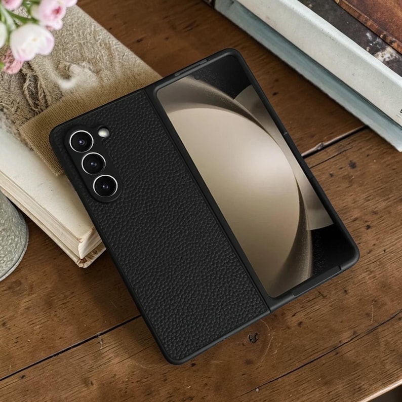 Puede incluir: Funda de tel&eacute;fono de cuero negro con una superficie texturizada, abierta para revelar la pantalla. La funda del tel&eacute;fono tiene tres lentes de c&aacute;mara. La funda del tel&eacute;fono est&aacute; sobre una superficie de madera.