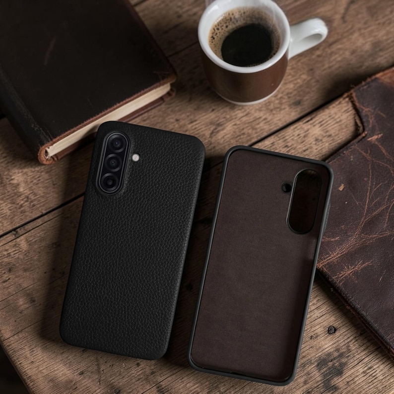 Op de afbeelding: Twee zwarte leren telefoonhoesjes, waarvan &eacute;&eacute;n open, liggen op een houten oppervlak naast een kopje koffie en een leren boek. De hoesjes hebben een getextureerde afwerking en een camera-uitsparing. De open hoes toont een bruin interieur.