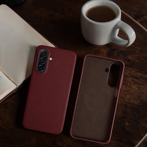 Op de afbeelding: Twee bordeauxrode leren telefoonhoesjes, waarvan &eacute;&eacute;n open, naast een witte koffiemok en een open boek op een donker houten oppervlak. De hoesjes hebben een getextureerde afwerking.