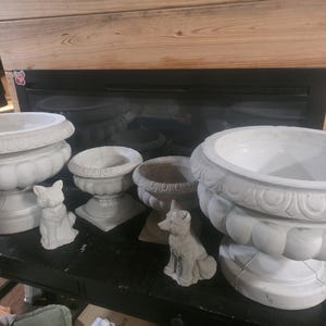 May include: A collection of white concrete planters and decorative animal figurines. The planters vary in size and design, with ornate detailing around the rims. Two small cat and fox figurines are also present, adding a whimsical touch to the display.