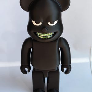 May include: A black vinyl figure with a bear-like design, featuring large ears and a stern expression. The figure's mouth is adorned with a set of sparkling, multi-colored gems. The toy stands upright, showcasing a modern, urban aesthetic.