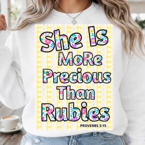 Può includere: Felpa bianca con la scritta "She Is More Precious Than Rubies" in un design floreale. Il testo è posto su uno sfondo a scacchi giallo e bianco con un bordo in pizzo. Il versetto biblico Proverbi 3:15 è in basso.