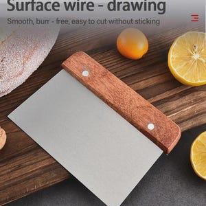 May include: A stainless steel bench scraper with a wooden handle. The text "Surface wire - drawing" is at the top of the image. The scraper is on a wooden surface with a loaf of bread, a lemon half, and a small orange.