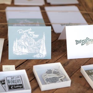 May include: A display of stationery items on a wooden surface. Includes greeting cards with text and illustrations, such as a mountain scene and the words "Renewed Day by Day." Also shown are stickers and small boxes.