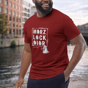 Könnte beinhalten: Ein weinrotes T-Shirt mit dem Text "MORZ LOCK IOR" in einem weißen Rahmen und einer Cartoon-Zeichnung eines rauchenden Huhns. Die Person trägt eine marineblaue Hose.