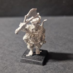 May include: A silver miniature figurine of a fantasy warrior, wielding a large axe. The figure has detailed fur-like textures on the shoulders and chest, and stands on a small, black rectangular base. The warrior is posed as if ready for combat.