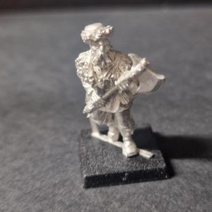 May include: A small metal figure of a person holding an axe. The figure is wearing a hat and a fur-like garment. The figure is standing on a small, black, rectangular base.