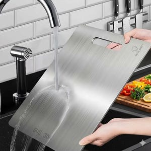 May include: A stainless steel chopping board with a handle is being washed under a tap. The board has the text "SUS 304" and the logo "METAL CHEFS". A wooden chopping board with chopped vegetables is in the background.