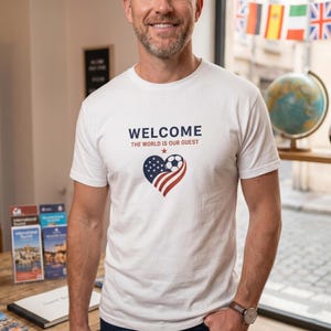 以下が含まれることがあります： 「WELCOME THE WORLD IS OUR GUEST」の文字と、アメリカ国旗とサッカーボールを組み合わせたハート型のデザインが施された白いTシャツ。半袖でクルーネックです。