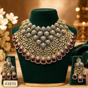 Puede incluir: Un conjunto de collar y pendientes dorados con intrincados detalles de filigrana y acentos de piedras preciosas rojas. El collar se exhibe sobre un busto verde, y los pendientes sobre un soporte a juego. El conjunto incluye el número #3870.