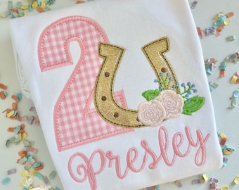 May include: A white onesie with a pink gingham number 2, a gold horseshoe with glitter, pink flowers, and the name "Presley" embroidered in pink.