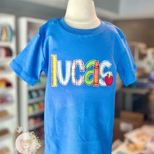 Puede incluir: Una camiseta azul de manga corta con el nombre "Lucas" en letras aplicadas de colores. El dise&ntilde;o incluye una regla, un l&aacute;piz de color y una manzana. La camiseta se muestra en un maniqu&iacute;.