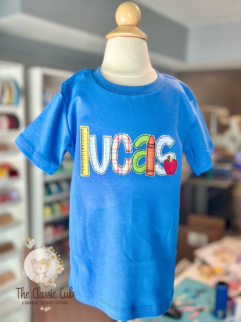 Puede incluir: Una camiseta azul de manga corta con el nombre "Lucas" en letras aplicadas de colores. El dise&ntilde;o incluye una regla, un l&aacute;piz de color y una manzana. La camiseta se muestra en un maniqu&iacute;.