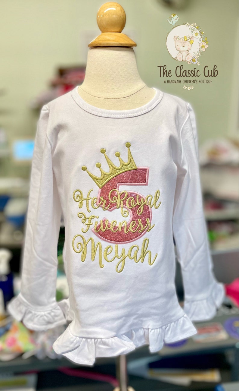 Peut inclure: Chemise blanche &agrave; manches longues avec une couronne dor&eacute;e et un chiffre 5 rose brod&eacute;s sur le devant. Le texte "Her Royal Fiveness Meyah" est &eacute;galement brod&eacute; en or.
