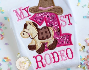 Peut inclure: Combinaison blanche brodée de rose et de marron. La combinaison dit "My 1st Rodeo" avec un numéro un rose, un chapeau de cowboy marron et un cheval marron.