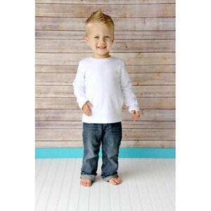 May include: A young boy wearing a white long-sleeved shirt and blue jeans stands in front of a wood panel background.