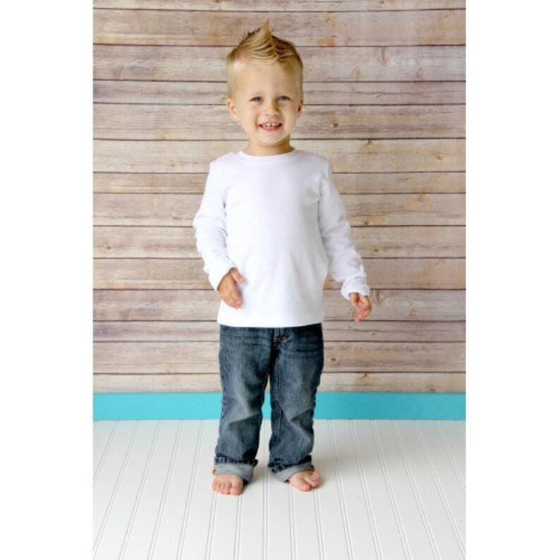 May include: A young boy wearing a white long-sleeved shirt and blue jeans stands in front of a wood panel background.