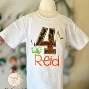 May include: White t-shirt with a camouflage number 4, a duck, and green reeds embroidered on it. The name "Reid" is embroidered in orange below the number.