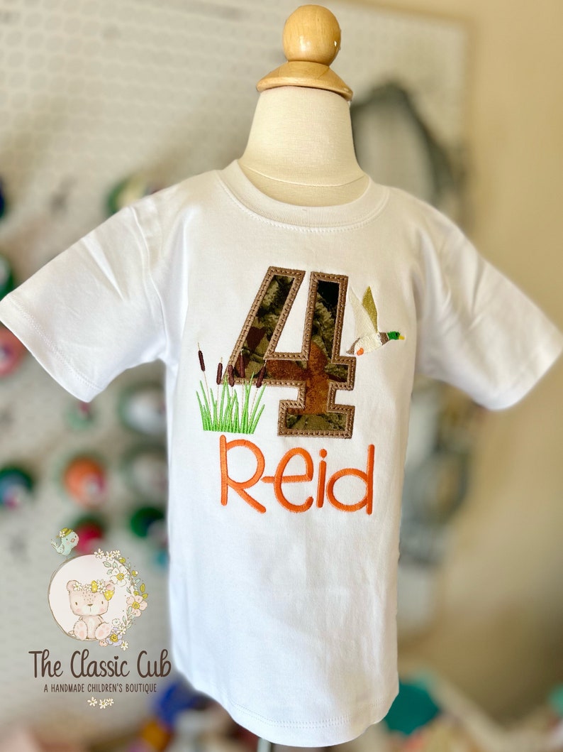 May include: White t-shirt with a camouflage number 4, a duck, and green reeds embroidered on it. The name "Reid" is embroidered in orange below the number.