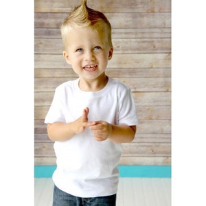 May include: A young boy with blonde hair styled in a mohawk is wearing a white t-shirt. He is smiling and looking at the camera.
