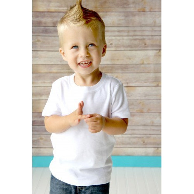 May include: A young boy with blonde hair styled in a mohawk is wearing a white t-shirt. He is smiling and looking at the camera.