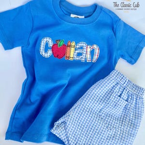 Puede incluir: Conjunto infantil azul compuesto por camiseta y pantal&oacute;n corto. La camiseta presenta el nombre "Collan" con un dise&ntilde;o de manzana y l&aacute;pices de colores. El pantal&oacute;n corto es de cuadros vichy azul claro y blanco.