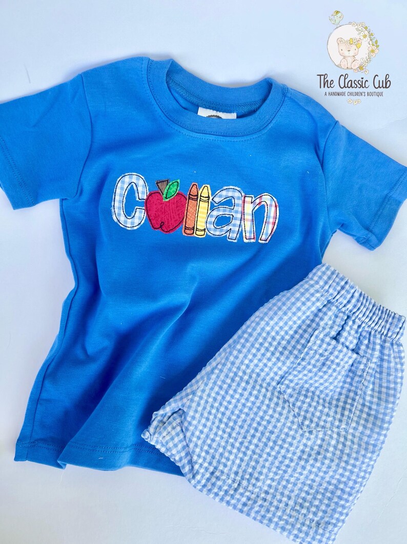 Puede incluir: Conjunto infantil azul compuesto por camiseta y pantal&oacute;n corto. La camiseta presenta el nombre "Collan" con un dise&ntilde;o de manzana y l&aacute;pices de colores. El pantal&oacute;n corto es de cuadros vichy azul claro y blanco.