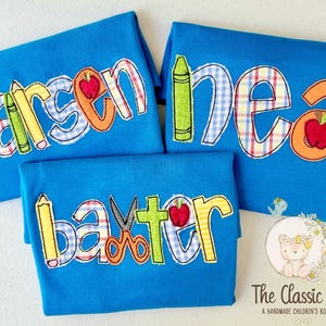 Puede incluir: Tres camisetas azules con nombres bordados y dise&ntilde;os escolares. Cada camiseta presenta un nombre diferente en letras coloridas, con &uacute;tiles escolares como crayones, manzanas y reglas.