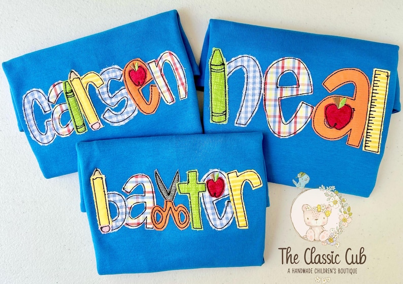 Puede incluir: Tres camisetas azules con nombres bordados y dise&ntilde;os escolares. Cada camiseta presenta un nombre diferente en letras coloridas, con &uacute;tiles escolares como crayones, manzanas y reglas.