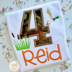 May include: A white t-shirt with a camouflage number four, a flying duck, and green grass. The name "Reid" is embroidered in orange below the number.