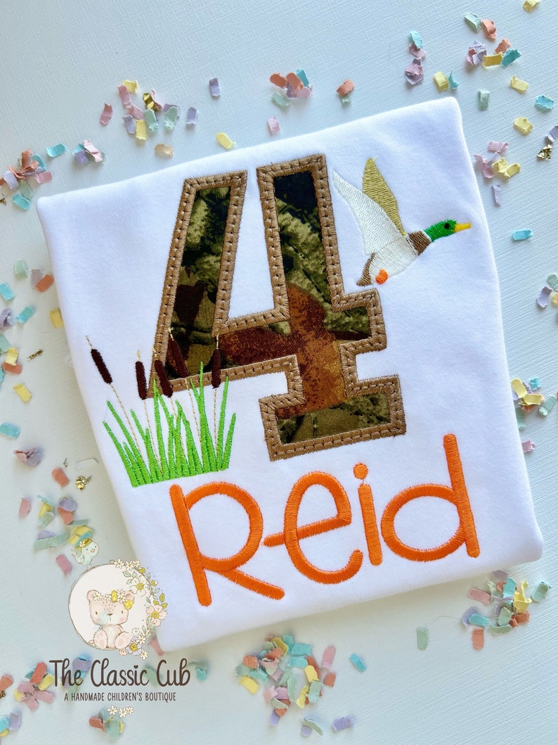 May include: A white t-shirt with a camouflage number four, a flying duck, and green grass. The name "Reid" is embroidered in orange below the number.