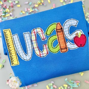 Puede incluir: Una camiseta azul con el nombre "LUCAS" en letras aplicadas coloridas. La "L" es una regla amarilla, la "U" tiene un patr&oacute;n de cuadros, la "C" es verde, la "A" tiene un l&aacute;piz naranja y la "S" una manzana roja.