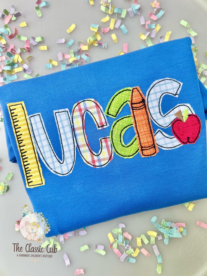 Puede incluir: Una camiseta azul con el nombre "LUCAS" en letras aplicadas coloridas. La "L" es una regla amarilla, la "U" tiene un patr&oacute;n de cuadros, la "C" es verde, la "A" tiene un l&aacute;piz naranja y la "S" una manzana roja.